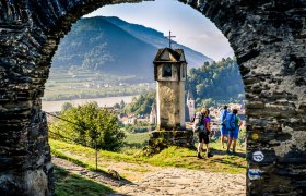 Aussicht vom Roten Tor in Spitz, © Robert Herbst Blick durch ein steinernes Tor auf eine Landschaft mit Hügeln, einem Fluss und Wanderern.