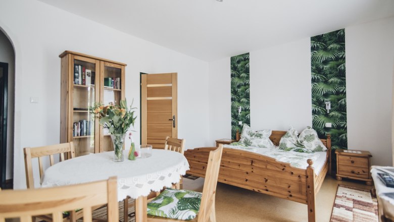 Doppelzimmer, © Józef Swierkot Ein gemütliches Doppelzimmer mit Holzmöbeln, einem Tisch mit Blumen, einem Bett mit Pflanzenmuster und einem Bücherregal.