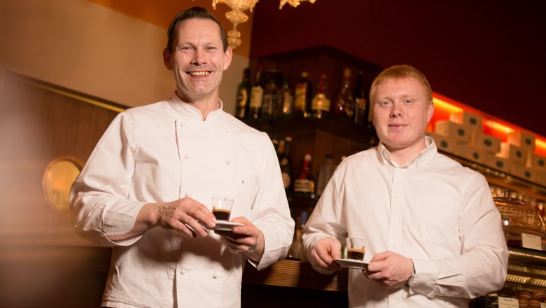 Cafe Helmut & Helmut, © schwarz-koenig.at Zwei Männer in einem Café halten Espressotassen und lächeln in die Kamera.