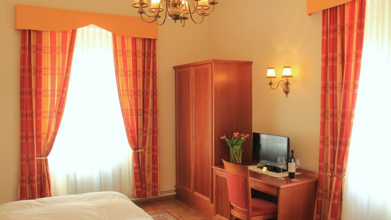 Doppelzimmer Superior, © Schendl Ein gemütliches Hotelzimmer mit Doppelbett, Holzmöbeln, einem Schreibtisch mit Fernseher und einer Vase mit Blumen. Vorhänge in Rot-Orange.