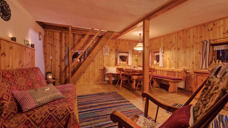 Wohn-Esszimmer, © Wachahof Gemütliches Wohnzimmer mit Holzwänden, Sofa, Schaukelstuhl und Esstisch. Rustikales Ambiente mit Teppich und warmem Licht.