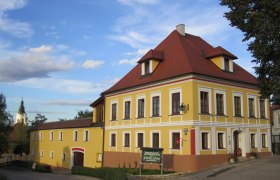 Hansy-Haus, © Fam. Schrammel Gelbes zweistöckiges Gebäude mit rotem Dach und weißen Fensterrahmen, daneben ein kleineres Gebäude. Im Hintergrund eine Kirchturmspitze.
