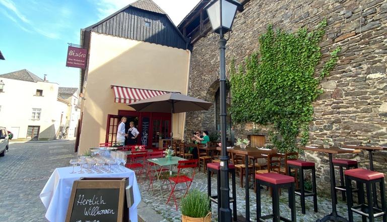 Bistro Krems, © Bistro Krems Ein gemütliches Bistro mit roten Stühlen und Tischen im Freien, umgeben von alten Steinmauern und Pflanzen.