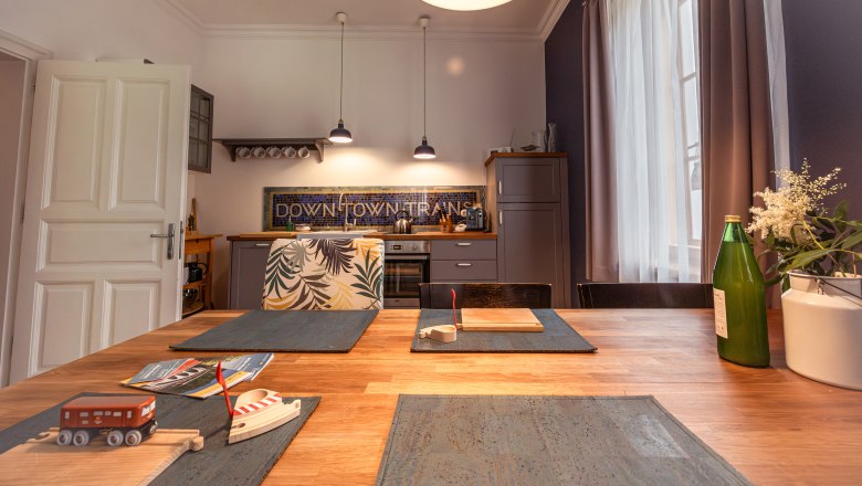 Urlaub am BAHNhof, © Niederösterreich Werbung / Maximilian Pawlikowsky Küche mit Holztisch, Spielzeugzug, Flasche und Blumen auf dem Tisch, moderne Lampen und Küchenschränke im Hintergrund.