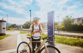 Am Bahnhof in Bad Erlach, © Wiener Alpen, Philipp Schönauer Eine Frau mit Fahrradhelm steht neben einem Fahrrad am Bahnhof in Bad Erlach.