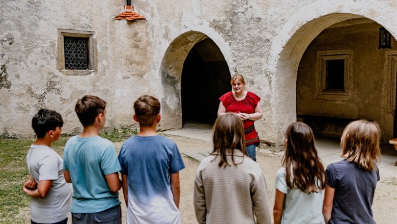 Burg Heidenreichstein, © Waldviertel Tourismus, Matthias Streibel Eine Frau in rotem Oberteil spricht zu einer Gruppe von Kindern vor einer alten Steinmauer mit Torbögen.