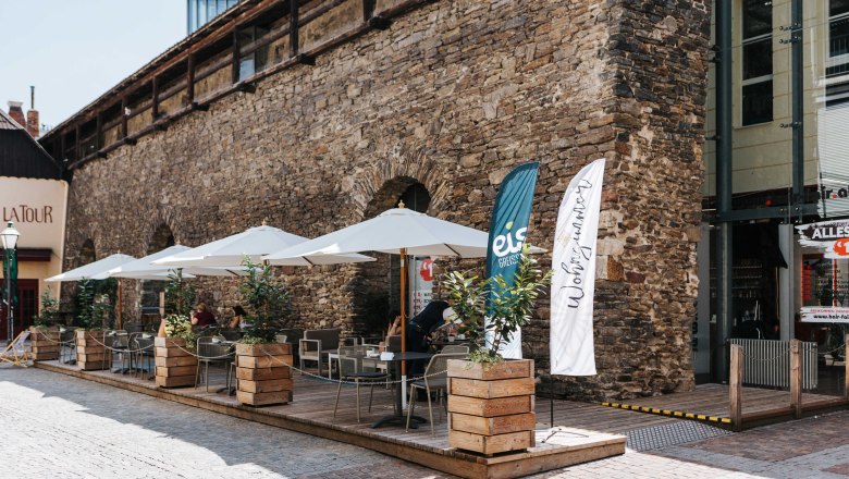 Café Wohnzimmer Gastgarten, © FMS Steinertor Gastronomie BetriebsgmbH Außenterrasse eines Cafés mit Sonnenschirmen und Pflanzen vor einer alten Steinmauer.