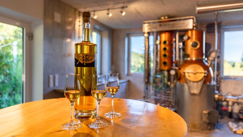 Brennanlage Familie Kölbel, © Wiener Alpen, Christian Kremsl Eine Flasche und drei halb gefüllte Gläser stehen auf einem runden Holztisch vor einer Brennanlage.