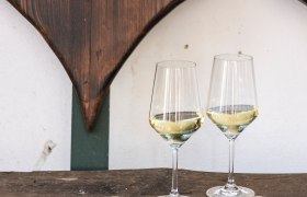 Weingläser, © Weinviertel Tourismus / Doris Schwarz-König Zwei Weingläser mit Weißwein auf einem Holztisch vor einer Holzherz-Dekoration.