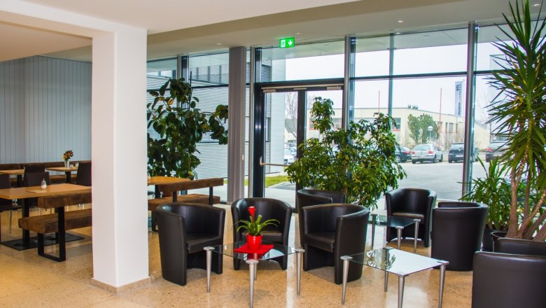 Cafe im Stadthotel, © Stadthotel Hollabrunn Innenansicht eines modernen Cafés mit schwarzen Ledersesseln und Glastischen, umgeben von großen Pflanzen und Fenstern.