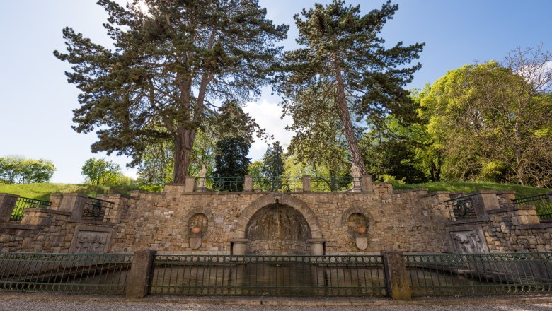 Großer Spuckmann_c_Schloss Artstetten/D. Mayrhofer, © Schloss Artstetten/D. Mayrhofer Steinmauer mit zwei großen Bäumen und dekorativen Elementen im Schlossgarten.