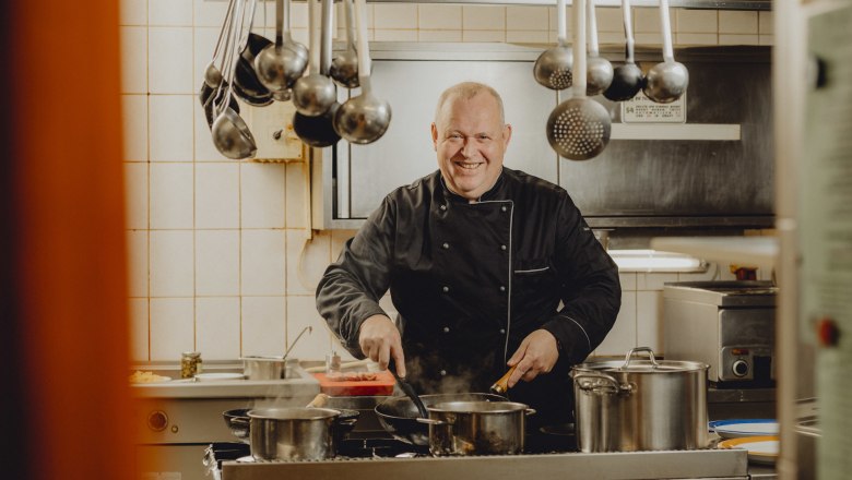 Chefkoch Jörg Hartl, © Niederösterreich Werbung/Rita Newman Ein Koch in schwarzer Uniform steht lächelnd in einer Küche und kocht an einem Herd mit mehreren Töpfen.