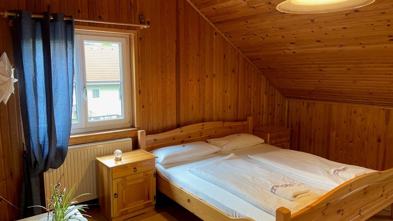 Doppelzimmer, © Wiener Alpen Gemütliches Doppelzimmer mit Holzverkleidung, Doppelbett, Fenster mit blauen Vorhängen und einer Pflanze auf dem Tisch.