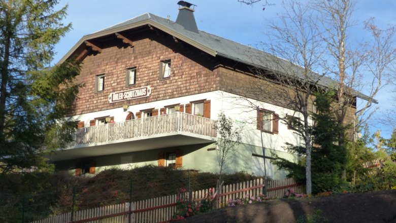 Die Schutzhütte, © Mario Putz Ein traditionelles Gebäude mit Holzverkleidung und Balkon, umgeben von Bäumen und einem Holzzaun. Schild: "Döhler-Schutzhaus".