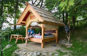 Bett im Freien in St. Corona am Wechsel, © Erika Kronaus Ein kleines Holzhaus mit einem Bett im Freien, umgeben von Bäumen. Zwei Personen sitzen und liegen auf dem Bett. Ein Picknicktisch steht daneben.
