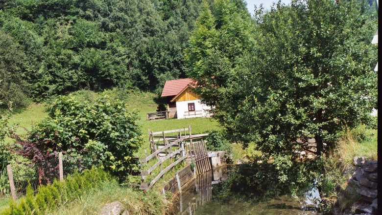 Pichlmühle, © Gemeinde Loich Eine kleine Hütte mit rotem Dach steht in einer grünen Landschaft neben einem Bach und Bäumen.