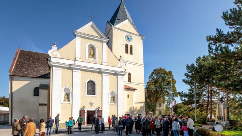 Kirchenplatz, © Gemeinde Prottes Menschen versammeln sich vor einer Kirche mit Turm und Uhr bei sonnigem Wetter.