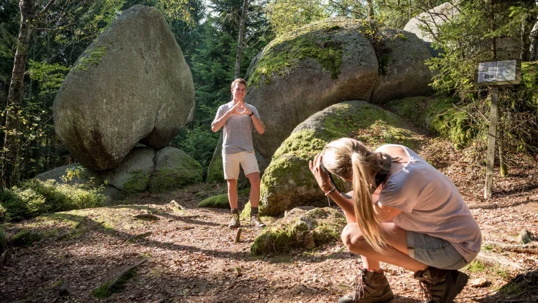 Herzstein, © Matthias Schickhofer Ein Mann posiert vor einem großen herzförmigen Felsen im Wald, während eine Frau ein Foto von ihm macht.