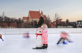 Eislaufplatz in Eggenburg, © Martin Mathes Ein Kind in pinker Kleidung steht auf einer Eisbahn mit einem Pinguin-Hilfsmittel. Im Hintergrund ist eine Kirche zu sehen.