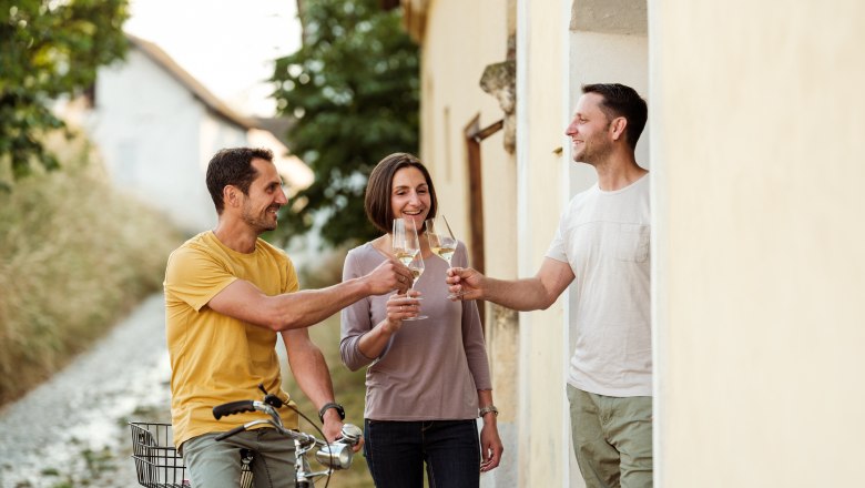 Geschwister Ewald, Maria und Christian, © Michael Reidinger Drei Personen stoßen mit Weingläsern an, eine Person sitzt auf einem Fahrrad.
