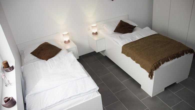 Arbeiterquartier UHL, © Arbeiterquartier UHL Zwei Einzelbetten in einem modernen, hellen Zimmer mit weißen Möbeln und braunen Decken.