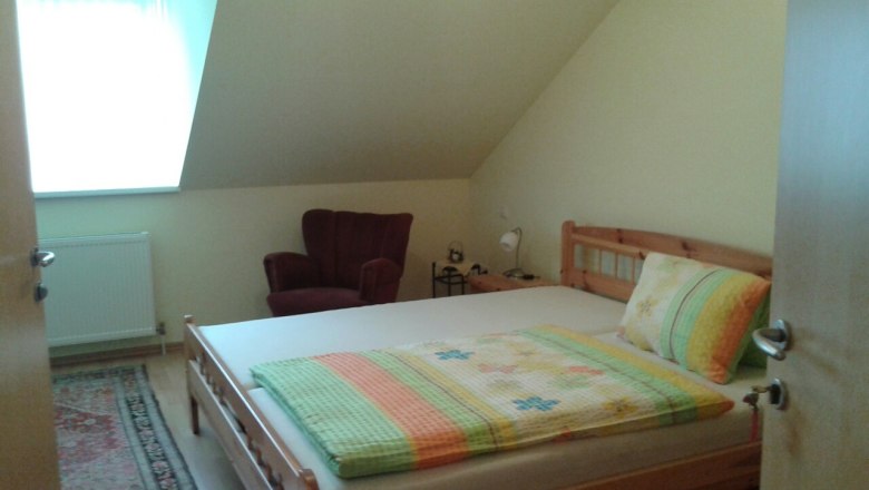 2. Doppelzimmer, © © Bianca Waysmayer Ein gemütliches Schlafzimmer mit einem Doppelbett, einem roten Sessel und einem kleinen Nachttisch unter einer Dachschräge.