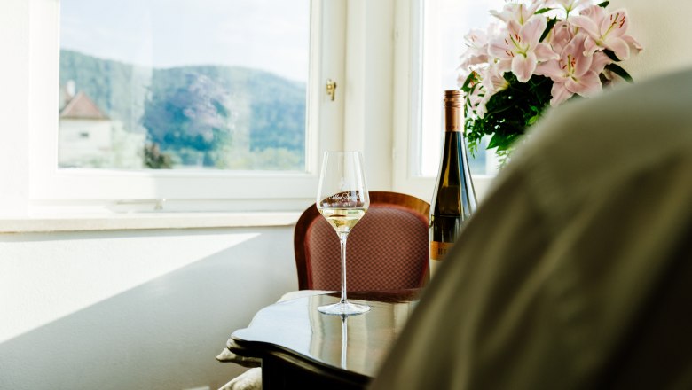 villa_schonthal-28_2, © Julius Hirtzberger Ein Weinglas und eine Weinflasche auf einem Tisch vor einem Fenster mit Blick auf Hügel und Blumen.