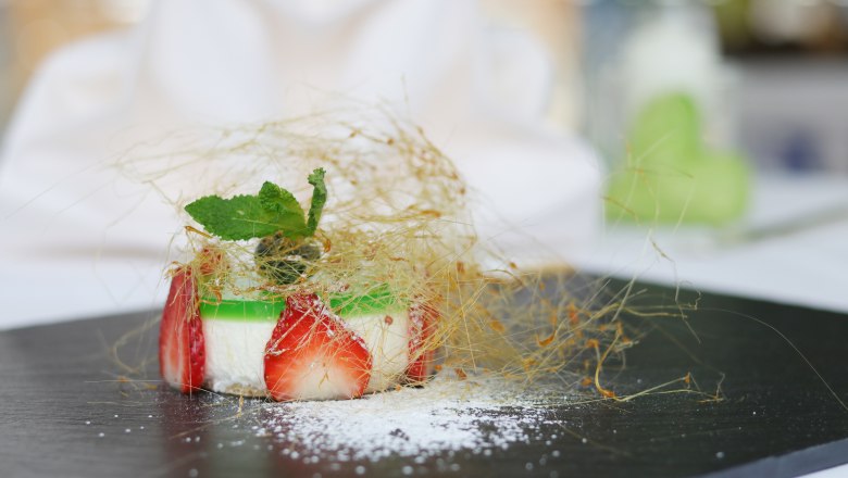 Haubenküche im Gesundheitsresort Königsberg: Ziegenkäsetörtchen, © Gesundheitsresort Königsberg Ein kunstvoll angerichtetes Ziegenkäsetörtchen mit Erdbeeren und Minze, umhüllt von Karamellfäden, auf einem schwarzen Teller.