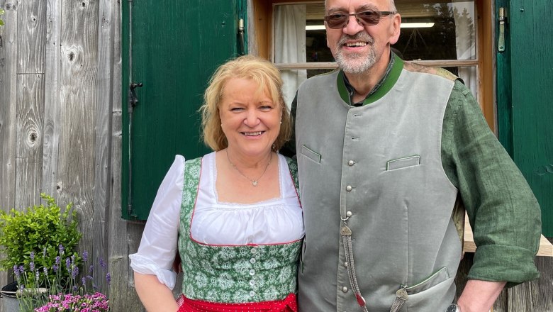 Familie Wetzelberger, © Lara Wetzelberger Ein Mann und eine Frau in traditioneller Tracht vor einem Holzhaus mit grünen Fensterläden.
