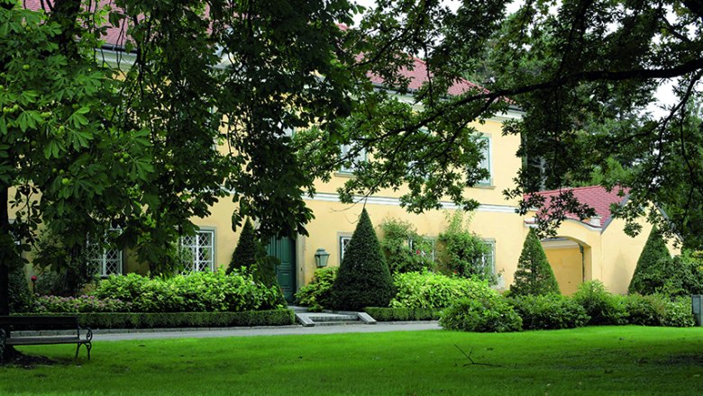 Parkanlage, © Gärtner Starkl Ein gelbes Gebäude mit roten Dächern, umgeben von grünen Bäumen und gepflegtem Rasen in einer Parkanlage.