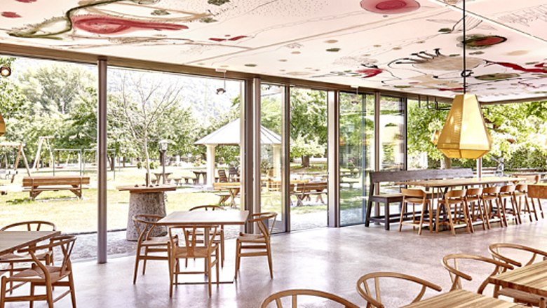 Innenansicht, © Familie Mang Innenansicht eines modernen Cafés mit großen Fenstern und Holzmöbeln, Blick auf einen grünen Außenbereich.