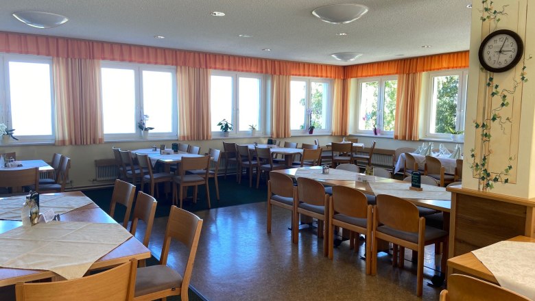 Speisesaal, © Wiener Alpen Ein heller Speisesaal mit Holzmöbeln, Tischen und Stühlen, dekoriert mit Pflanzen und einer Wanduhr.