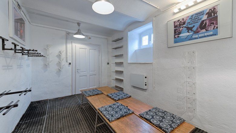 Skikeller, © VP FeWo OG Ein Skikeller mit weißen Wänden, Skihalterungen, einer Holzbank mit Kissen und einem Filmplakat an der Wand.