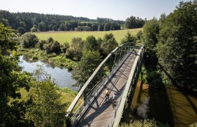 Thayarunde, Radfahren, © Waldviertel Tourismus, Erwin Haiden Die sanfte Brücke schwingt über den glitzernden Fluss, umgeben von üppigem Grün und der Ruhe der Natur. Radfahrer genießen die frische Luft und die malerische Landschaft, während die Sonne sanft auf die Wiesen scheint.