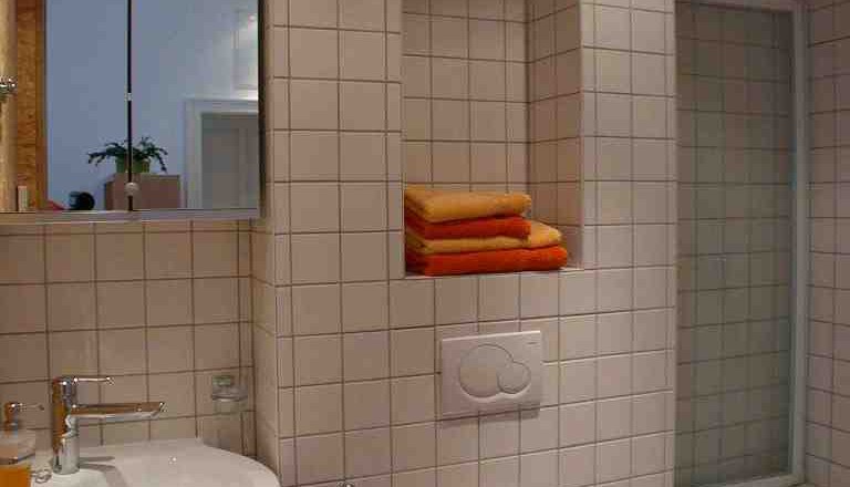 Badezimmer, Apartment Gutmann, © Gutmann Modernes Badezimmer mit weißen Fliesen, Waschbecken, Toilette und Badewanne. Handtücher in einem Regal.