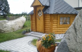 Grillhütte, © Grill Manfred Eine kleine, hölzerne Grillhütte mit grauem Dach, umgeben von Steinen und Pflanzen.