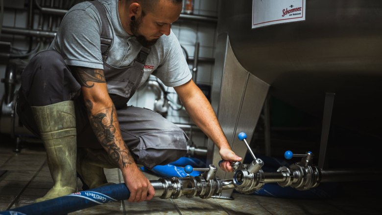 Brauen, © Inge Funke Ein Mann in Arbeitskleidung hält Biergläser vor einer Brauerei.