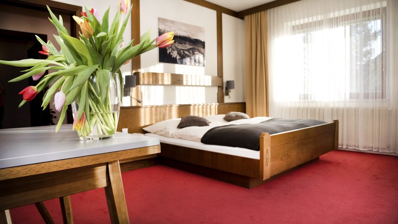 Doppelzimmer, © Doris Seebacher Ein gemütliches Doppelzimmer mit einem Holzbett, roten Teppichboden und einem Tisch mit einem Blumenstrauß aus Tulpen.
