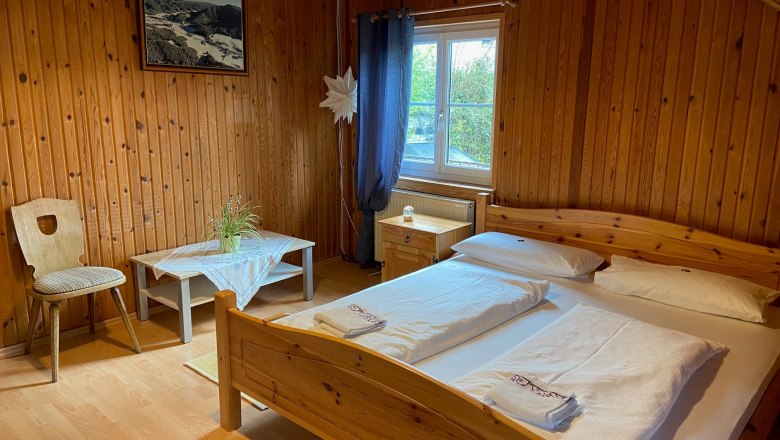 Doppelzimmer, © Wiener Alpen Holzvertäfeltes Doppelzimmer mit Bett, Tisch, Stuhl und Fenster.