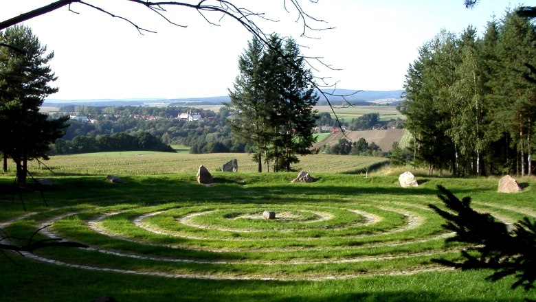 Arena Geomantica, © Naturpark Dobersberg Ein spiralförmiges Labyrinth aus Gras und Steinen auf einer Wiese mit Bäumen und einem Dorf im Hintergrund.