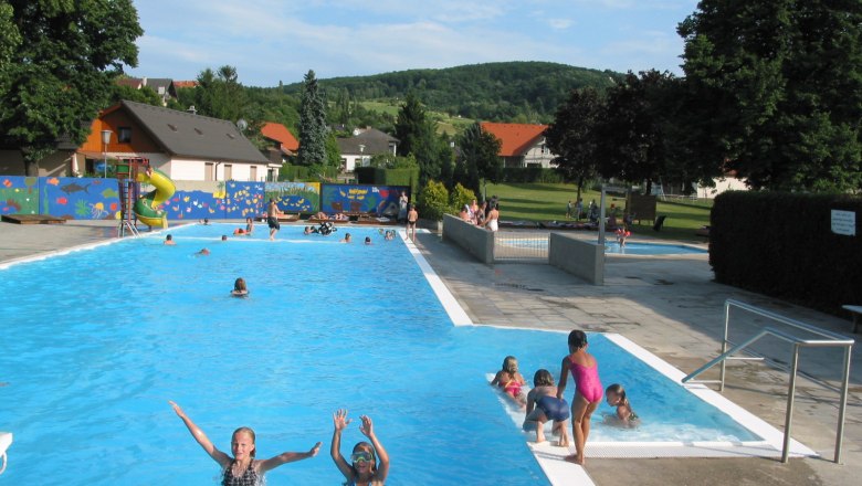 Freibad Königstetten, © Donau Niederösterreich Freibad mit Schwimmern und Rutsche, umgeben von Bäumen und Wiese.