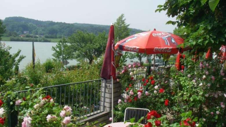 Garten, © Wachauerhof Ein blühender Garten mit roten und rosa Rosen, einem Tisch mit Stühlen und einem roten Sonnenschirm mit Coca-Cola-Logo, im Hintergrund ein See.