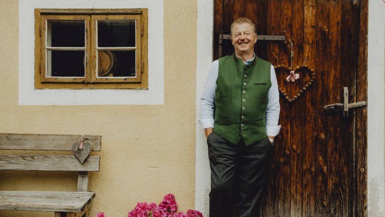 Wirt Anton Schilling, © Niederösterreich Werbung/Rita Newman Ein Mann in traditioneller Kleidung steht lächelnd vor einer Holztür neben einem Fenster und einer Bank mit Blumen.