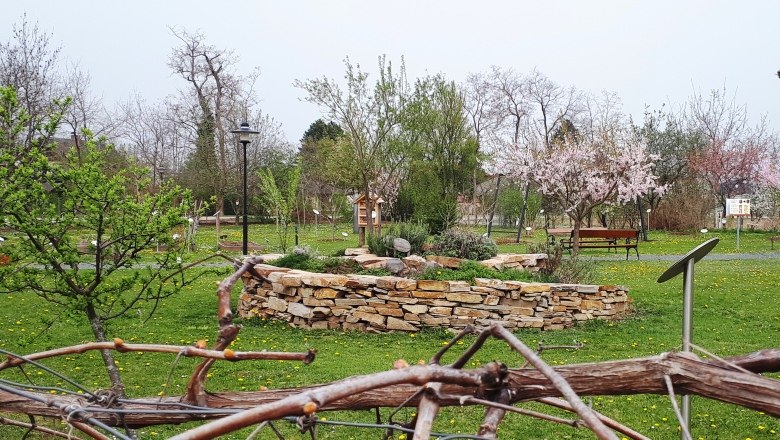 Kräuterspirale Alchemistenpark, © "Natur im Garten" Schaugärten Kräuterspirale im Alchemistenpark mit blühenden Bäumen und grüner Wiese.