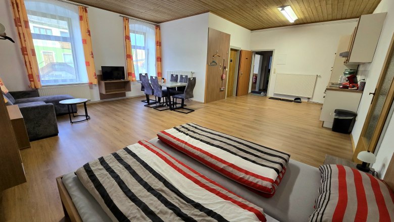 Aparment 4 - Schlafbereich mit Küche, © Apartment Bauer Zur Alten Schule Innenansicht eines geräumigen Apartments mit Holzboden, Doppelbett, Esstisch, Sofa und Fernseher.