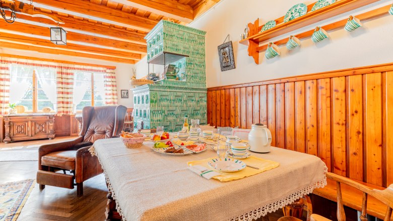 Herrenhaus Woltron, © Wiener Alpen / Christian Kremsl Gemütliches Esszimmer mit Holzmöbeln, gedecktem Tisch und grünem Kachelofen.