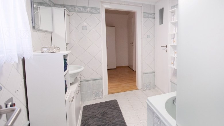 Badezimmer, © Ferienhaus Scheer Modernes Badezimmer mit weißen Fliesen, Badewanne, Waschbecken und Schrank. Tür führt zu einem Flur mit Holzboden.