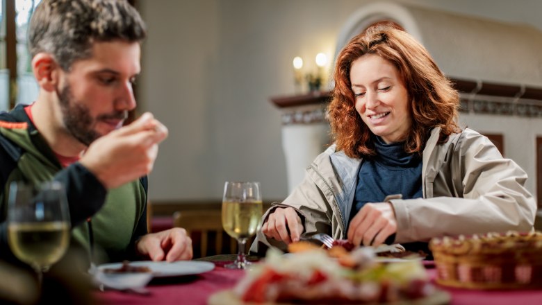 Beim Genussbauernhof und Heurigen Böhm in Katzelsdorf, © Wiener Alpen/Fülöp, Kremsl Ein Mann und eine Frau sitzen in einer Stube auf einem rustikalen Holztisch und essen eine Brettljause