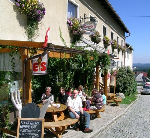 Gasthof zum Wachtstein, © Marktgemeind Bad Traunstein Eine Gruppe von Menschen sitzt vor einem Gasthof an Holztischen auf einer gepflasterten Straße.