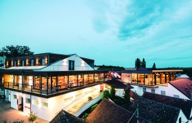 Weindomizil Hagn, © Weingut Hagn / Michael Reidinger Moderne Weingut-Architektur bei Dämmerung mit beleuchteten Innenräumen und umliegenden Gebäuden.